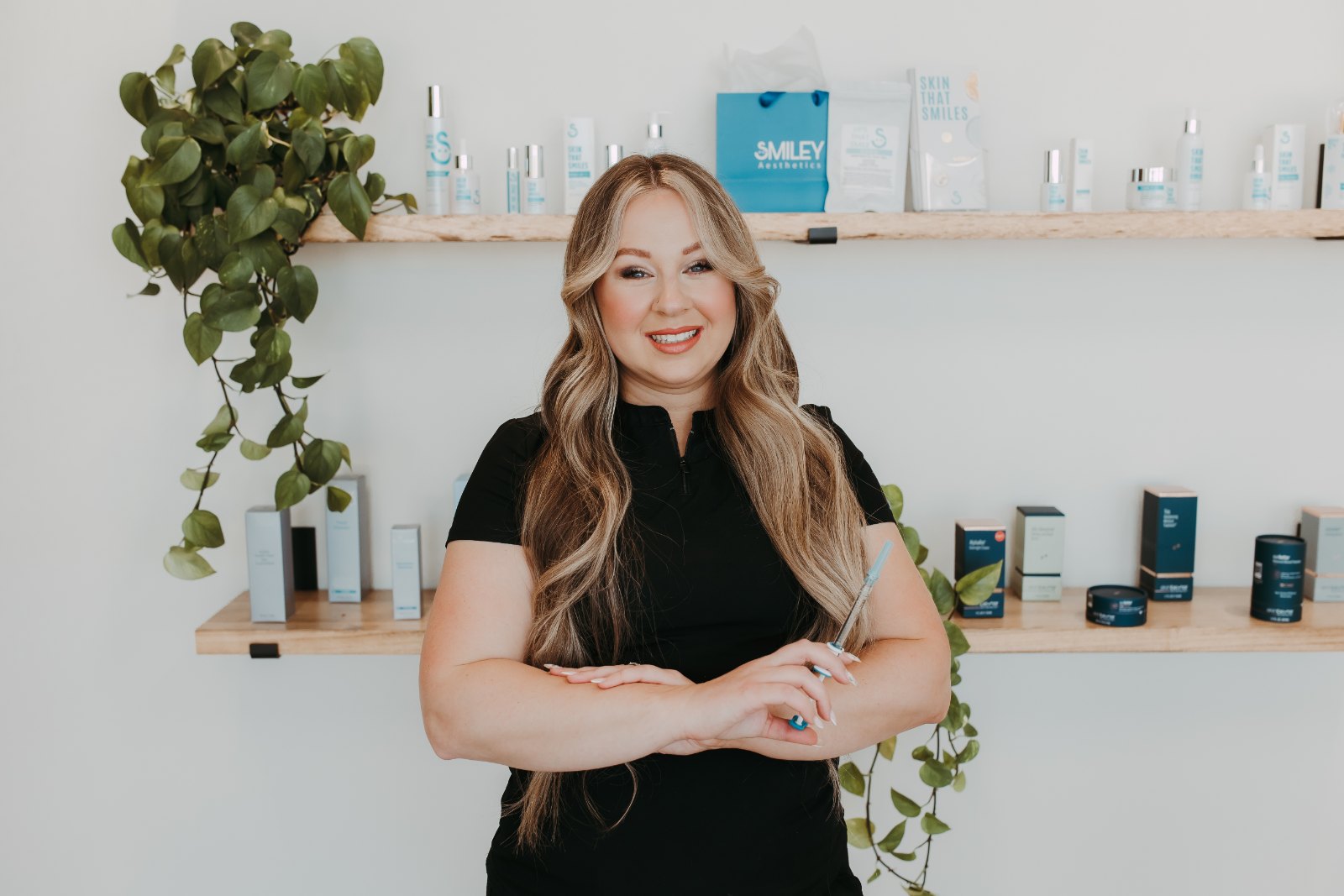 A Smiley Aesthetics provider in black scrubs smiling in front of a shelf of skincare products